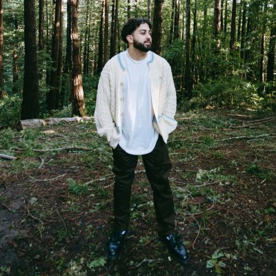 Man with dark hair and beard standing in a forest wearing a white oversized t-shirt, cream knitted cardigan, black pants, and black shoes.