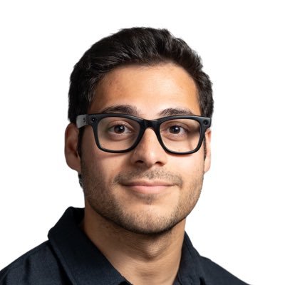 Portrait of a young man with short dark hair, beard, black-framed glasses, and a black collared shirt against a white background.