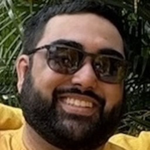 Portrait of a young man with short dark hair, beard, black-framed glasses, and a black collared shirt against a white background.