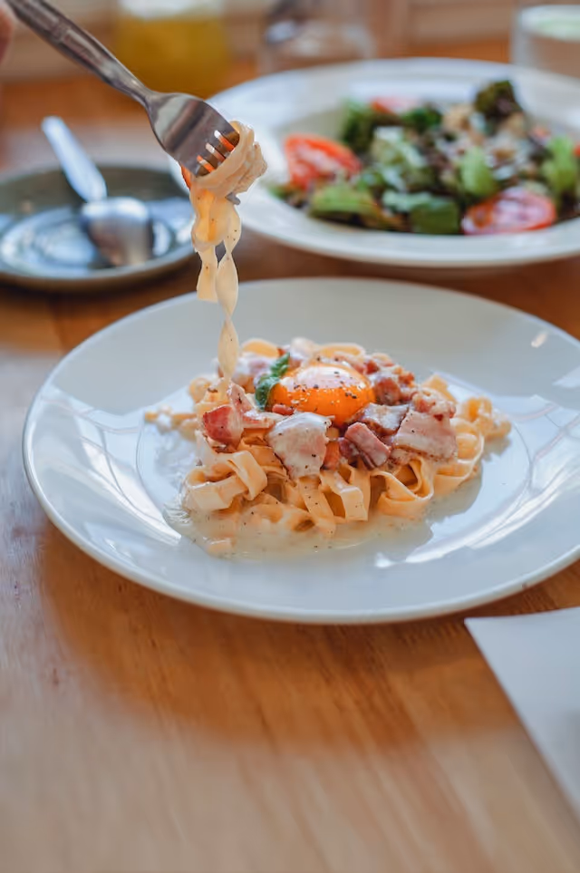 A plate of carbonara pasta with a fork, alongside a fresh salad, as someone prepares to take a bite.