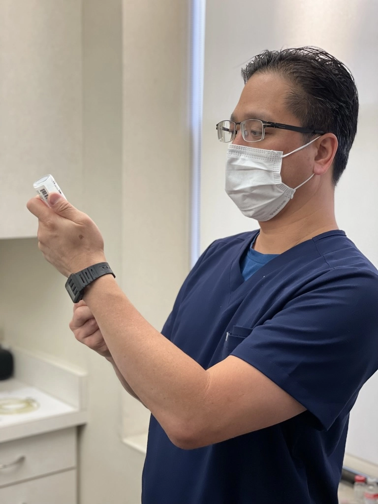 A male healthcare professional wearing a mask, glasses, and navy scrubs examines a medication vial in a clinical setting.