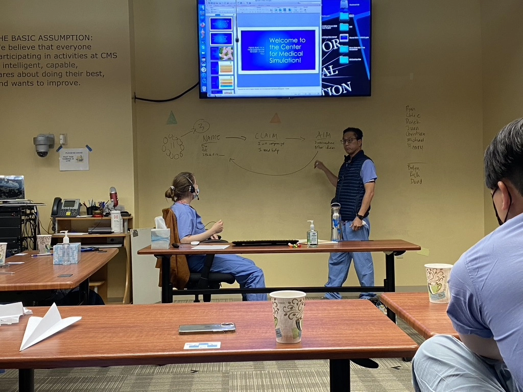 Dr. Richard Marn leads a training session at the Center for Medical Simulation, standing near a whiteboard and screen while participants in scrubs listen attentively.