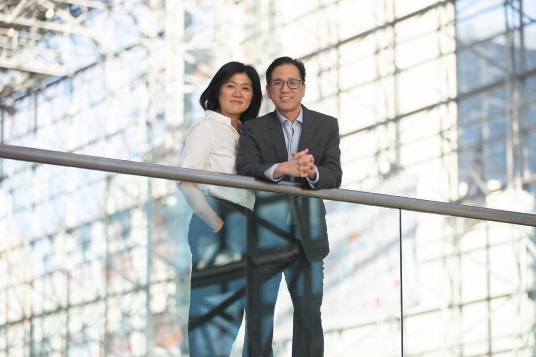 A smiling couple poses together in a modern glass building, with the man in a suit and the woman in a white blouse and dark pants.
