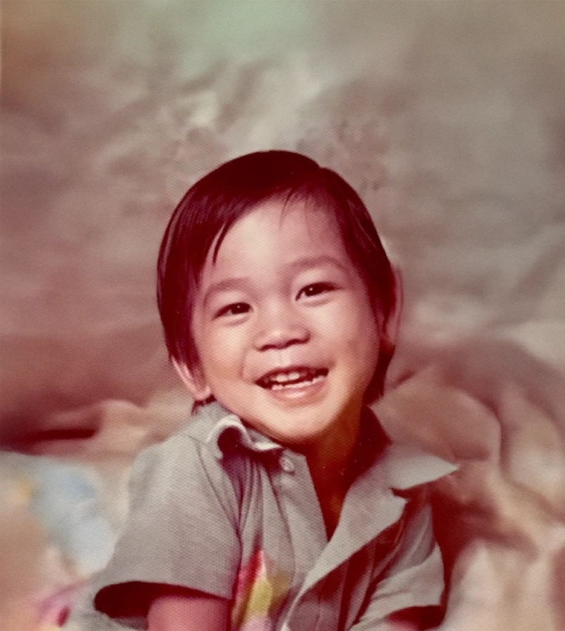 A young child with a joyful smile poses for a studio portrait, wearing a short-sleeved shirt with a soft-focus background.