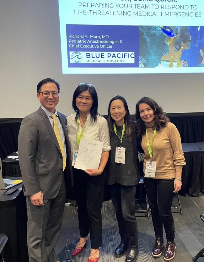 Dr. Richard Y. Marn stands with his wife and two women at a medical conference, with a Blue Pacific Medical Simulation presentation displayed in the background.