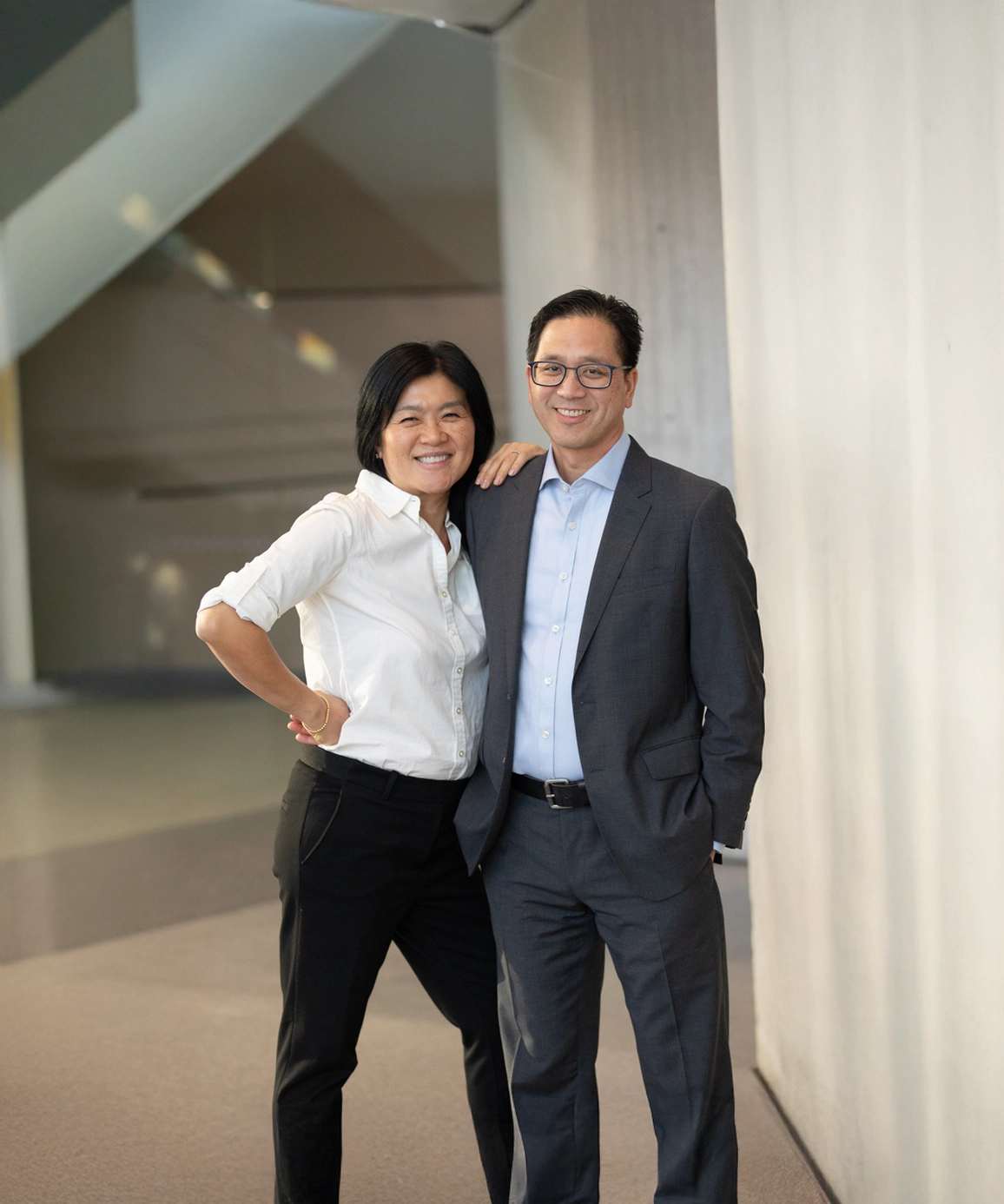 A smiling couple poses confidently in a modern, well-lit indoor setting; the man wears a gray suit and the woman wears a white blouse with black pants.