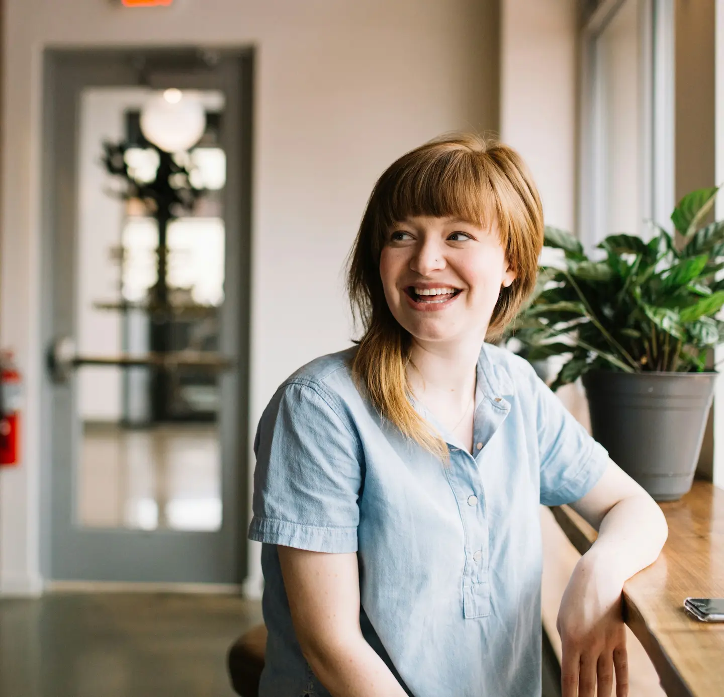 Smiling woman with red hair sitting at a wooden counter next to a potted plant in a bright room.