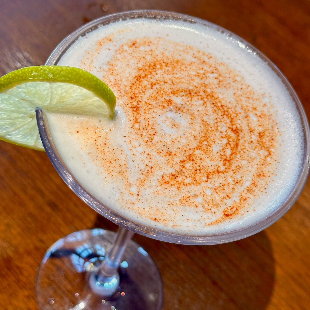 Cocktail in a stemmed glass with frothy top sprinkled with reddish spice and garnished with a lime wedge.