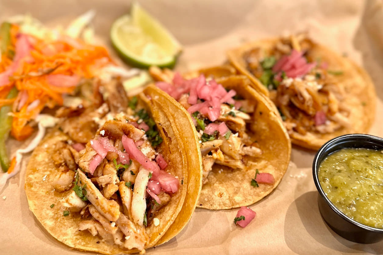 Three grilled chicken tacos topped with chopped onions and cilantro, served with lime wedge, pickled vegetables, and green salsa.