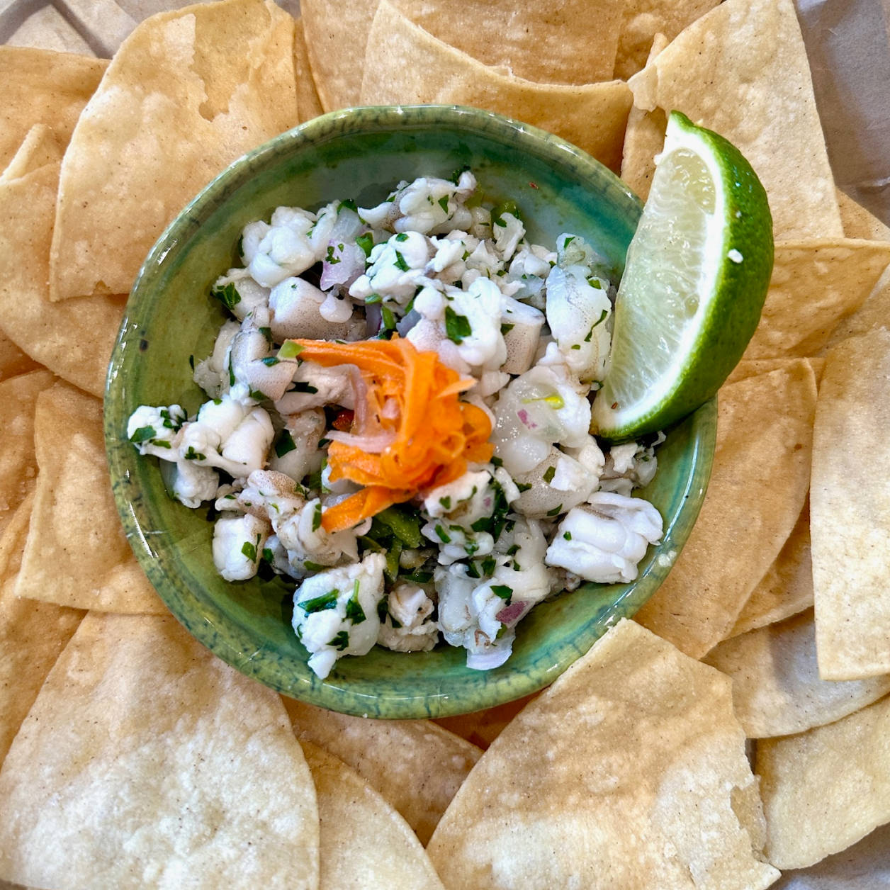 Green bowl of seafood ceviche garnished with shredded carrots and lime wedge, surrounded by tortilla chips.