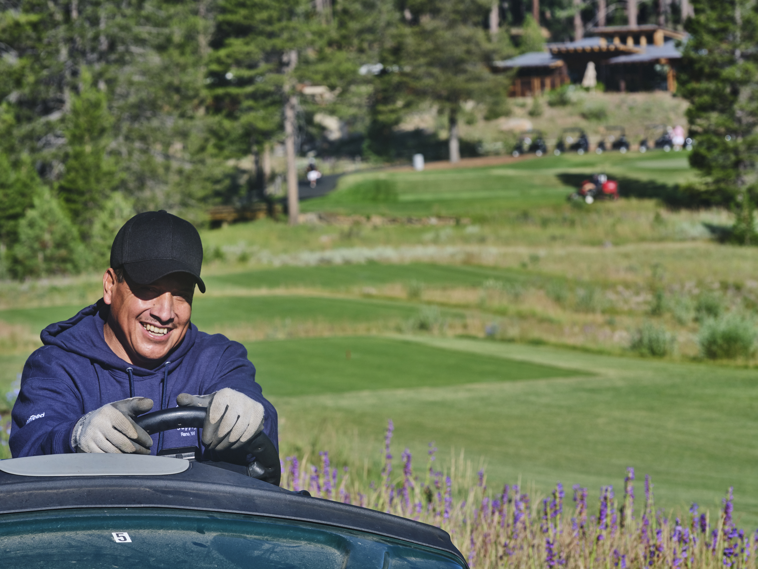 Smiling man wearing gloves and a black cap driving a golf cart on a green golf course with purple flowers in the foreground.