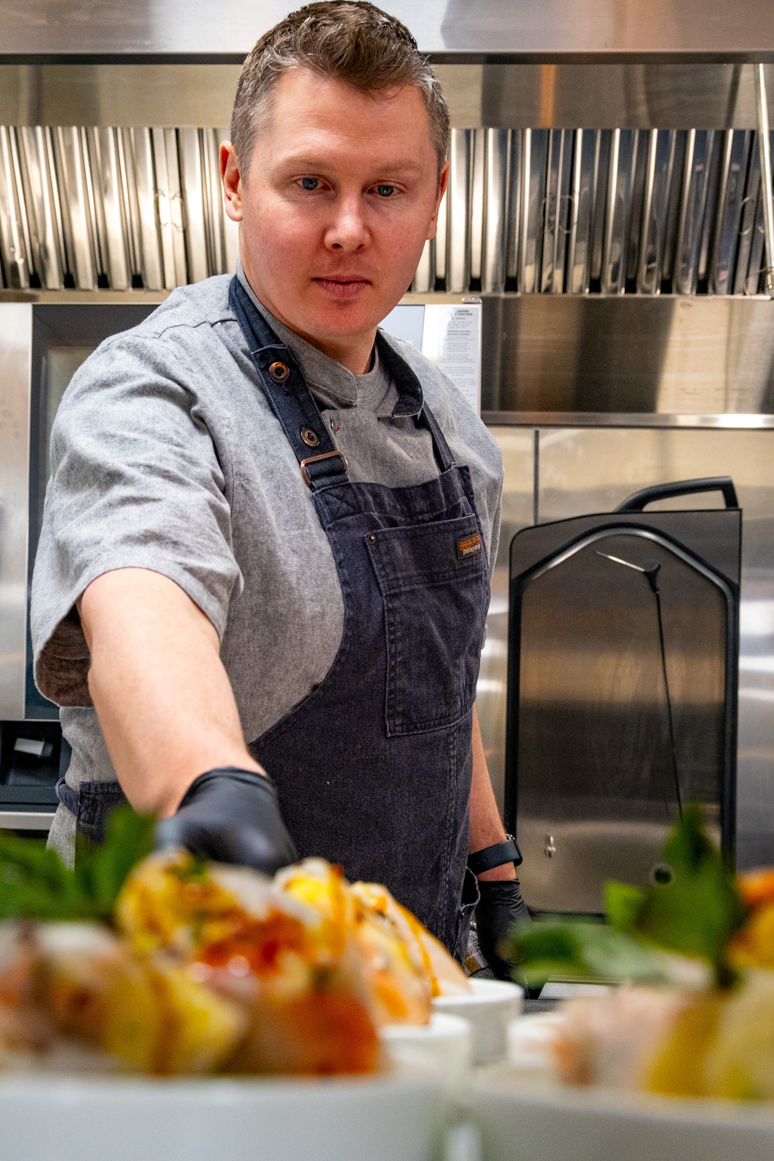 Chef in a gray shirt and dark apron arranging plated dishes in a professional kitchen.