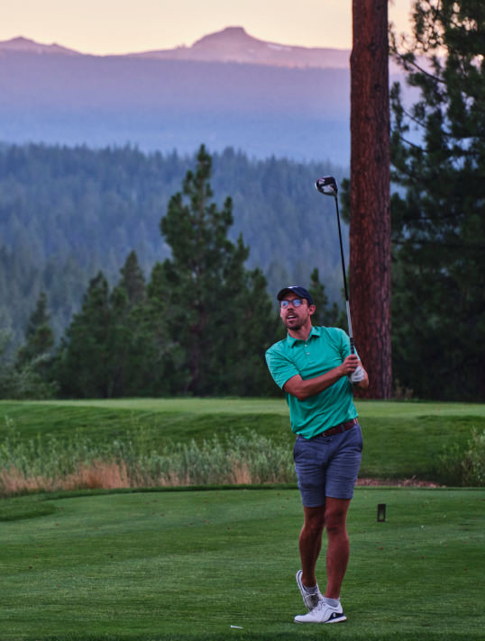Man in green polo shirt and shorts swinging a golf club on a golf course with forested mountains in the background at sunset.