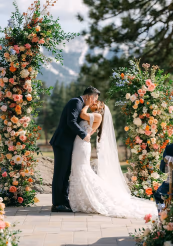 Bride and groom kissing under floral arch during outdoor wedding ceremony with trees and mountains in the background.