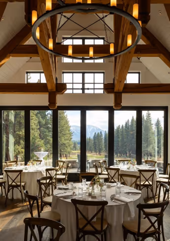 Indoor dining area with round tables set for meals, wooden chairs, large windows showing a forest and mountain view, and exposed wooden ceiling beams with a circular light fixture.