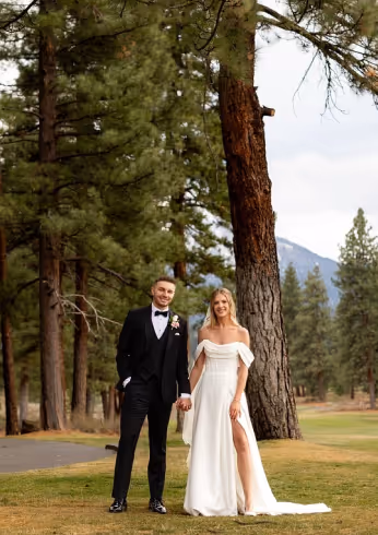 Bride in white off-shoulder gown and groom in black tuxedo standing hand in hand outdoors with pine trees and mountains in the background.