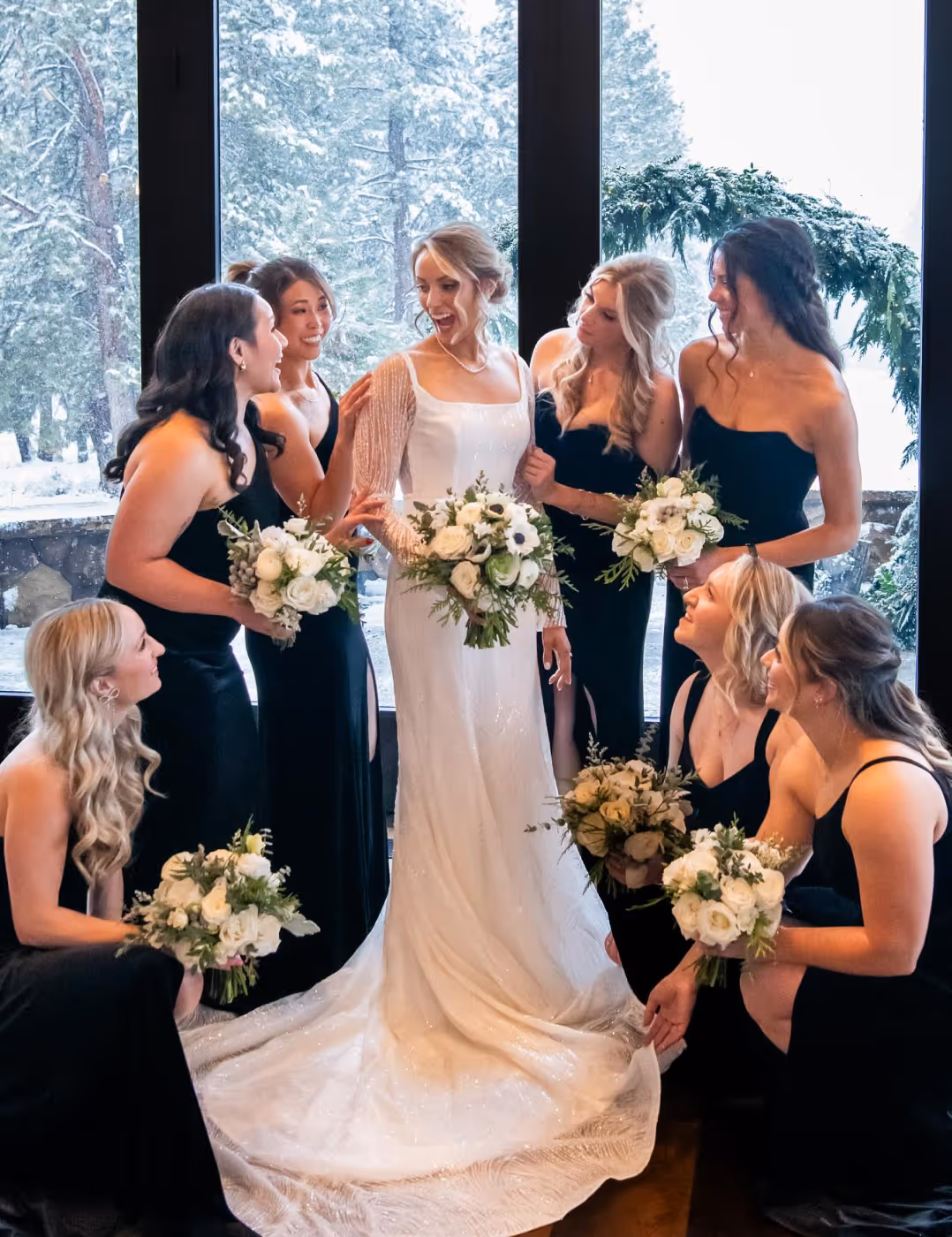 Bride in a white wedding gown surrounded by seven bridesmaids in black dresses holding white floral bouquets, with snowy trees visible through large windows behind them.