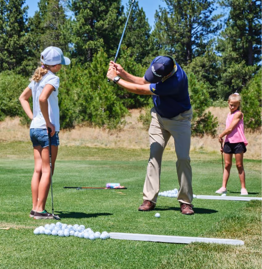 Man demonstrating a golf swing to two young girls on a golf course driving range with many golf balls on the grass.