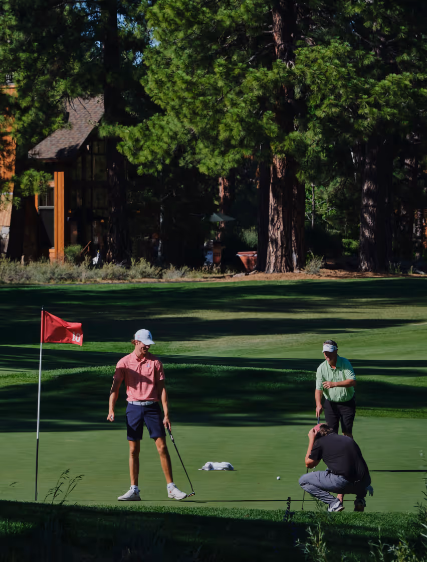 Three men playing golf on a green near a hole with a red flag numbered 10, surrounded by tall trees and a building in the background.