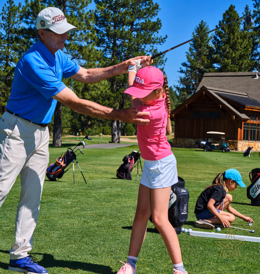 Golf instructor guiding a young girl’s swing on a sunny golf course while another child practices with golf balls nearby.