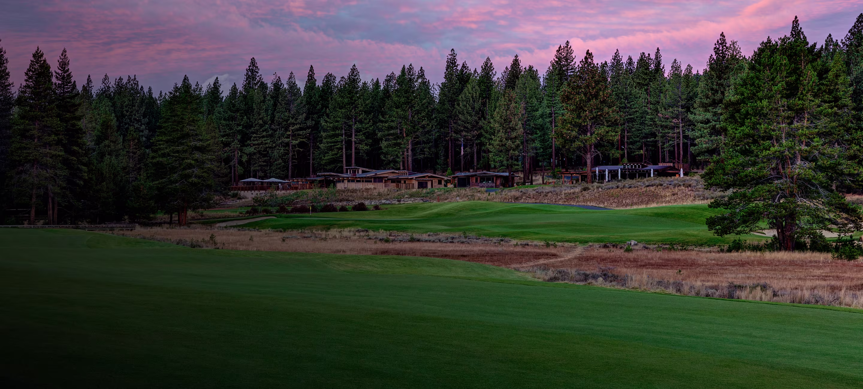 Golf course fairway and green with pine trees and cabins under a purple and pink sunset sky.
