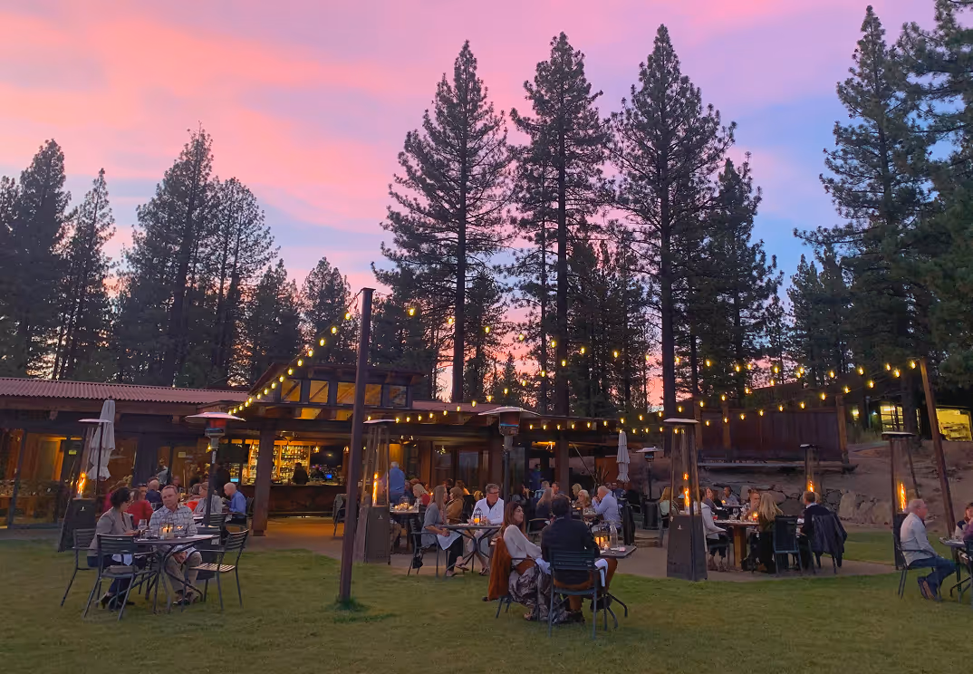 Outdoor dining area at sunset with people sitting at tables under string lights surrounded by tall pine trees.