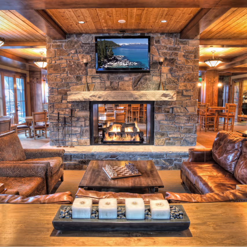 Cozy lodge interior with a stone fireplace, burning fire, wooden furniture, leather sofas, and a chessboard on a wooden table.