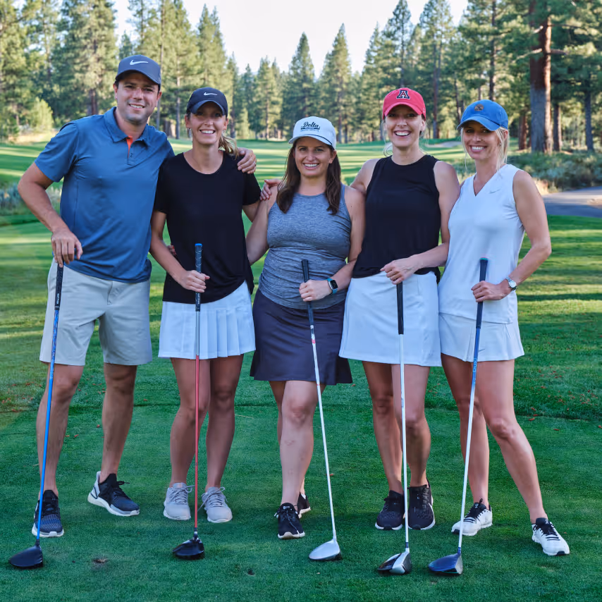 Five smiling golfers standing on a green golf course, each holding a golf club, with trees in the background.