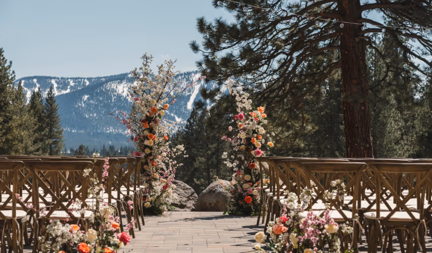 Outdoor wedding aisle decorated with colorful flowers and wooden chairs, set against a backdrop of pine trees and snow-capped mountains.