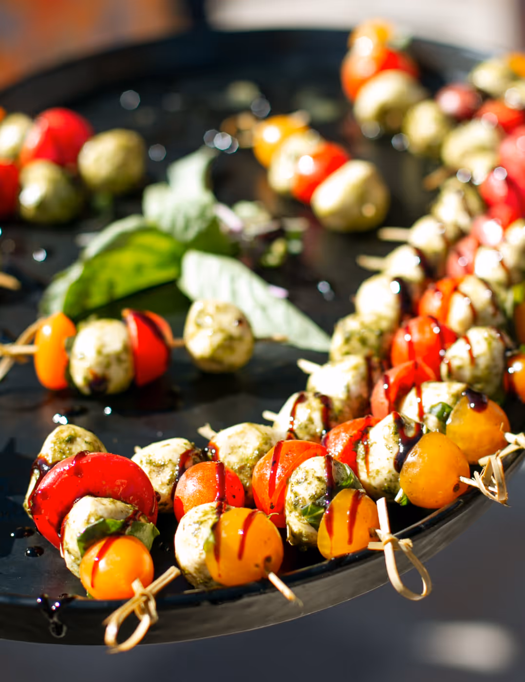 Skewers with cherry tomatoes, mozzarella balls, basil, and balsamic glaze on a black platter.