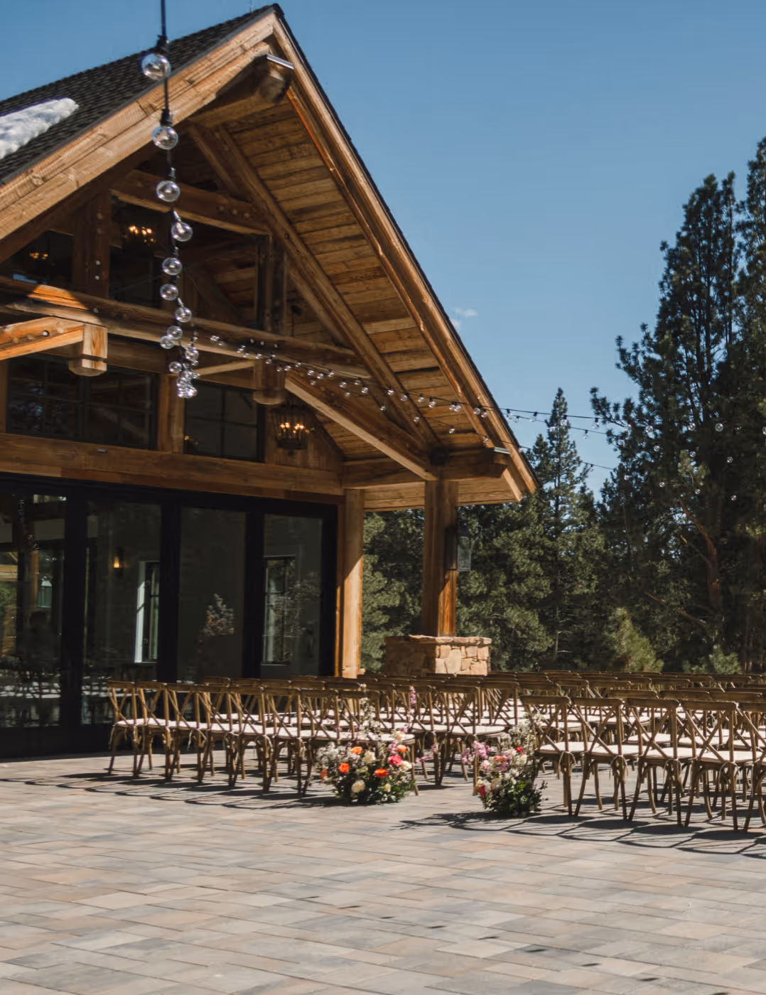 Outdoor event space with wooden chairs arranged in rows on a stone patio next to a rustic wooden building under a clear blue sky.