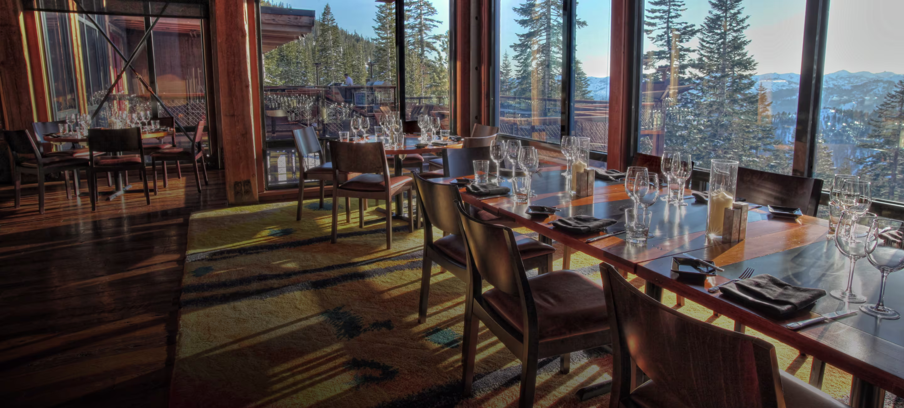Sunlit restaurant dining area with wooden tables and chairs set with wine glasses, overlooking snow-covered mountains through large windows.