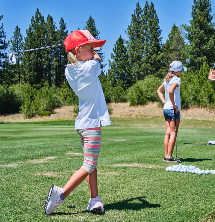 Young girl wearing a red cap swinging a golf club on a green golf course with another girl and golf balls in the background.