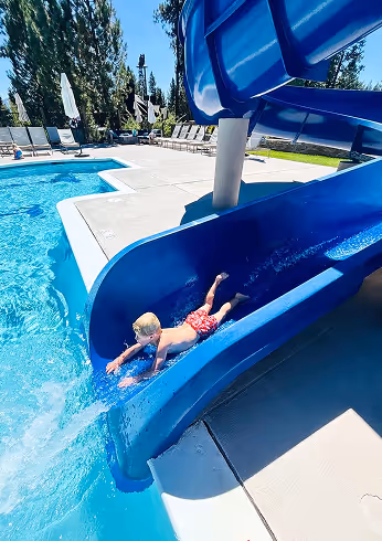 A young boy in red swim trunks slides headfirst down a blue water slide into a swimming pool on a sunny day.