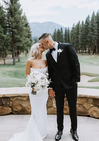 Bride in a white strapless dress holding a bouquet and groom in a black tuxedo kissing outdoors with trees and mountains in the background.