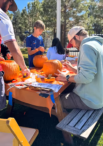 People carving pumpkins at a wooden picnic table outdoors on a sunny day.