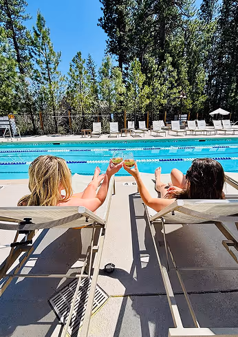 Two women lounging on pool chairs by an outdoor swimming pool, clinking glasses under a clear blue sky with trees in the background.