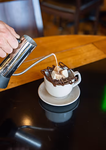 Hand pouring cream from a metal pitcher into a cup of coffee with a chocolate and marshmallow lattice on top.