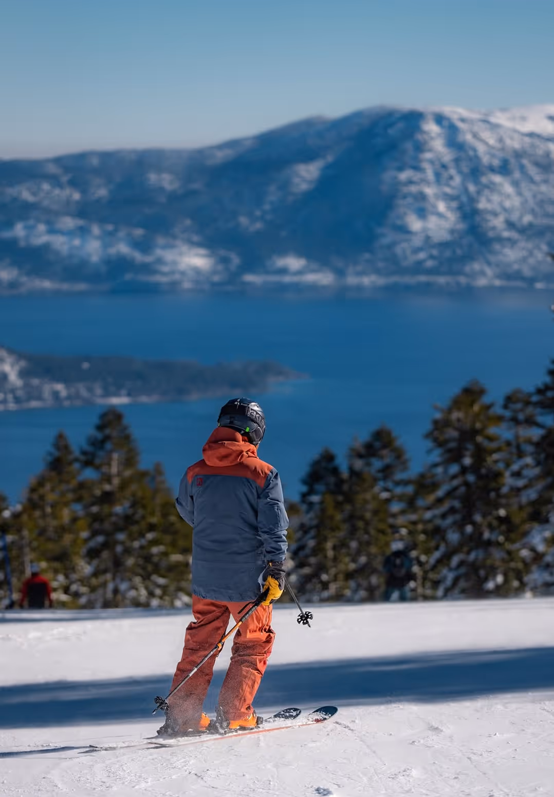 Person in orange and gray ski outfit standing on snow with ski poles, overlooking a lake and snow-covered mountains.