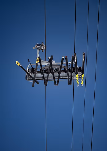 Skiers sitting on a chairlift suspended in the air against a clear blue sky with skis attached to the lift's footrest.