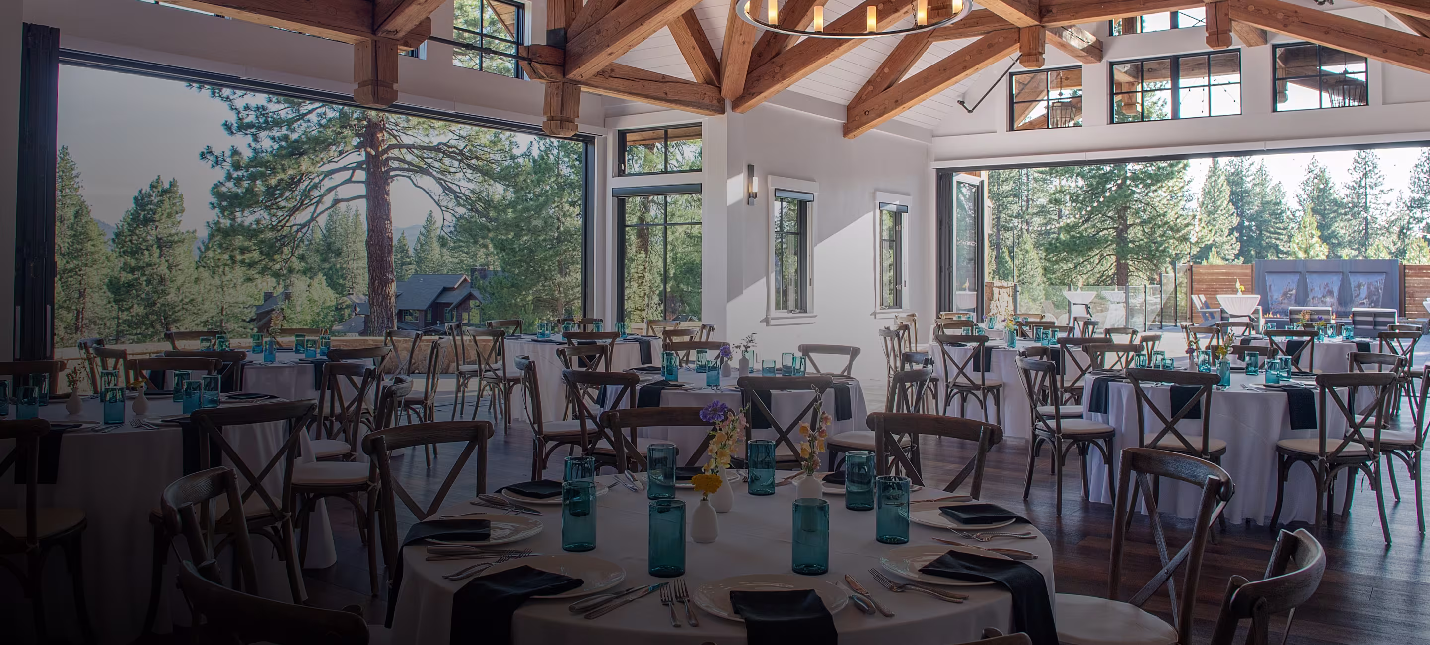 Spacious dining room with round tables set with blue glasses, black napkins, white plates, and small floral centerpieces, featuring large windows showing a pine tree forest outside.