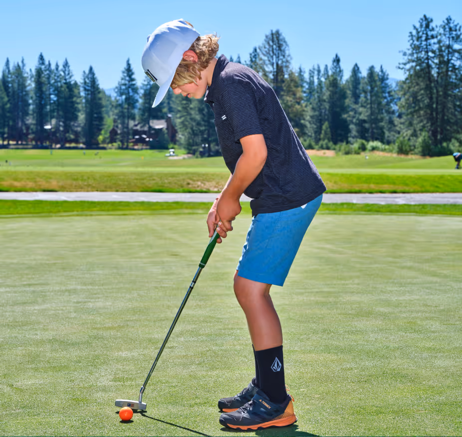 Boy wearing a white cap putting an orange golf ball on a green golf course on a sunny day.
