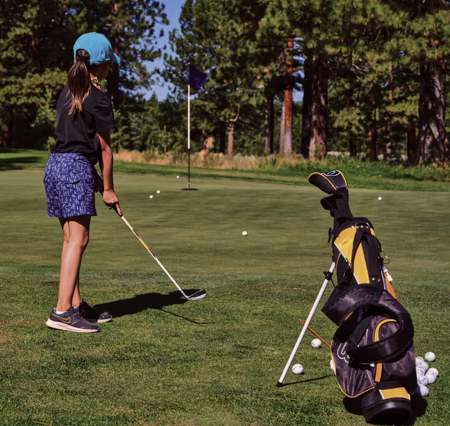 Child wearing a blue cap putting a golf ball on a green near a flag with golf bag and balls nearby.