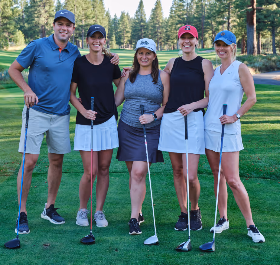 Group of five golfers standing on grass with golf clubs and trees in background.