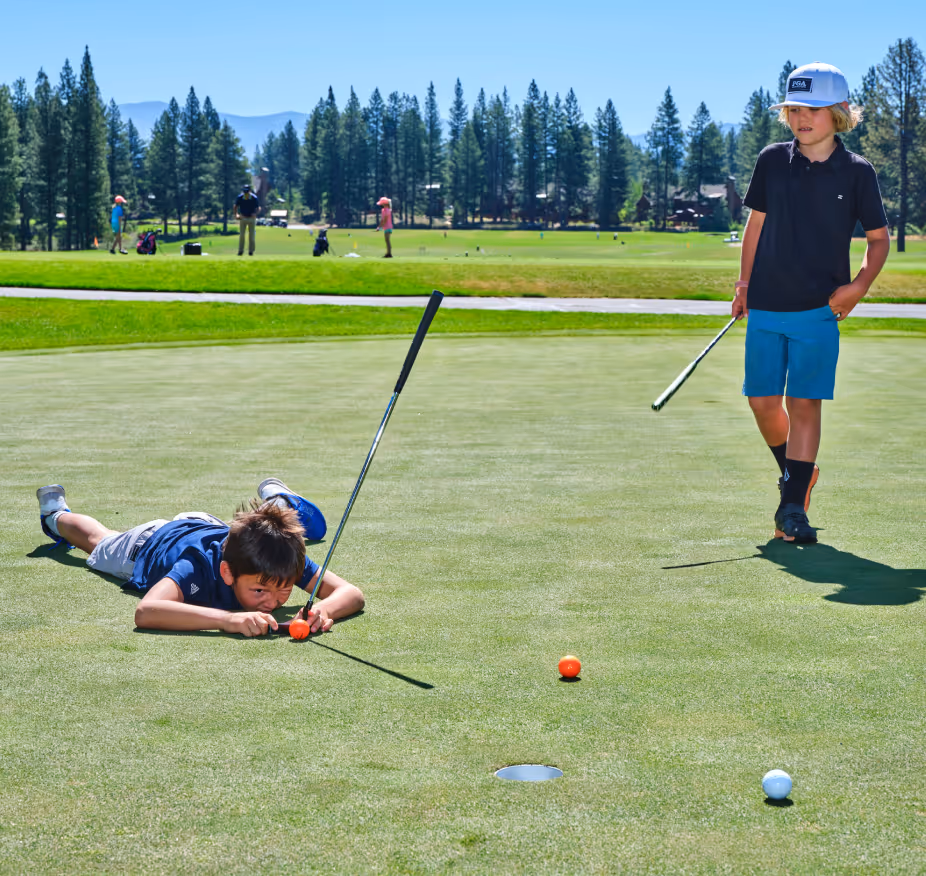 Two boys on a golf green, one lying down lining up a putt, the other standing holding a putter.