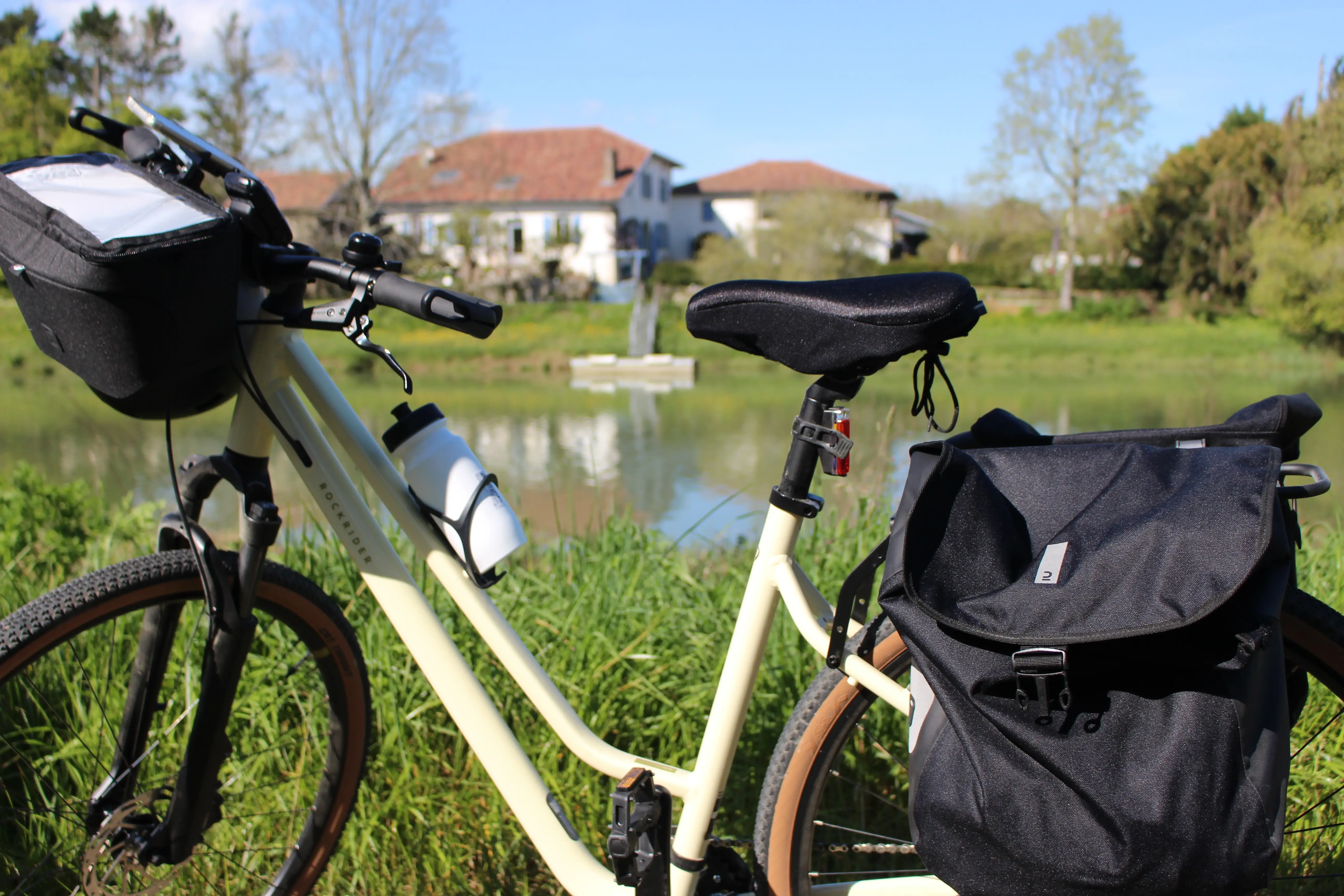 Vélo de randonnée blanc crème avec sacoches noires et bouteille d'eau posé devant un étang avec des maisons et des arbres en arrière-plan.
Location de vélo de voyage avec l'agence The Family Bike.