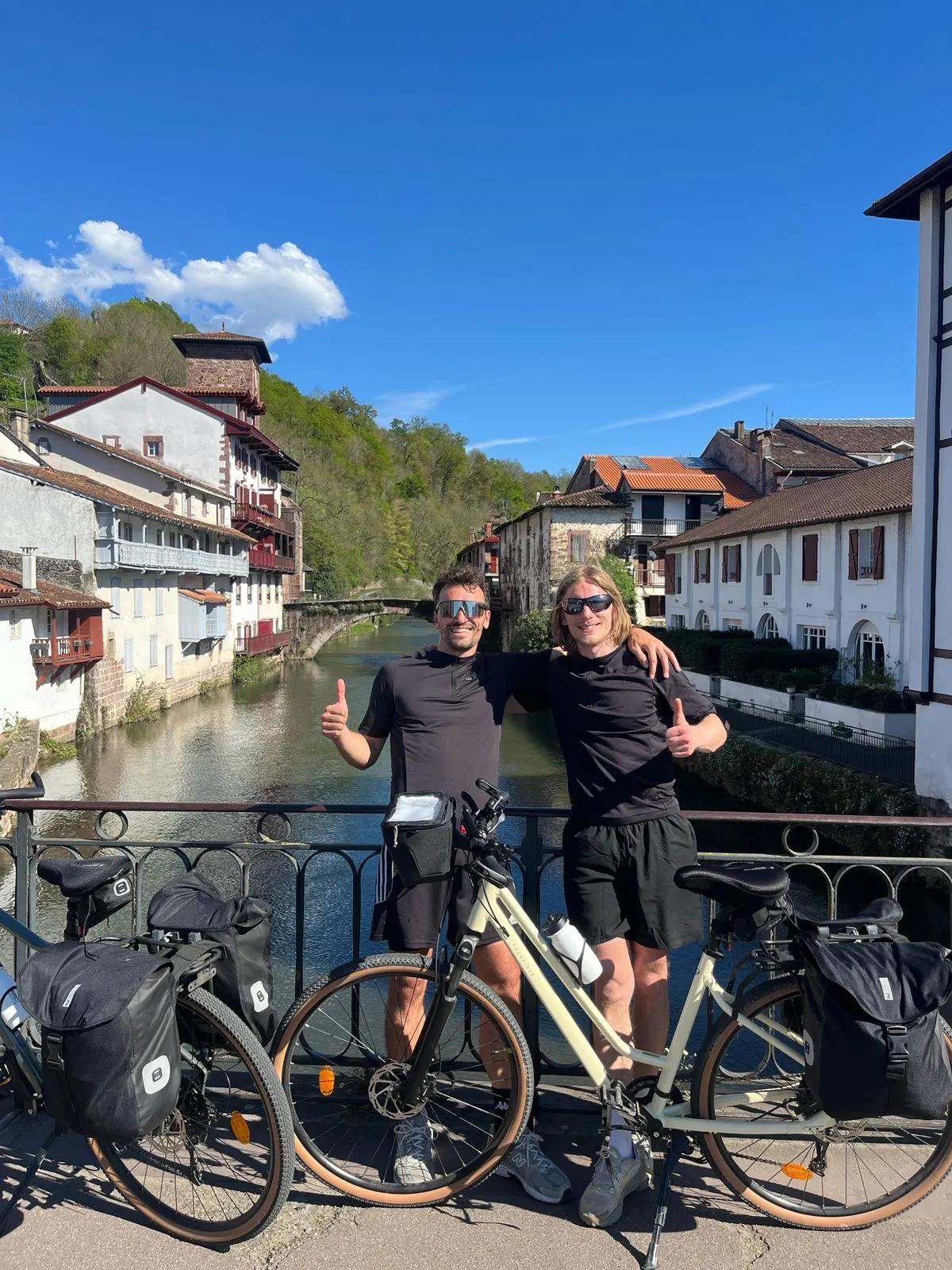 Deux hommes en tenue de cyclisme posent avec leurs vélos sur un pont à Saint Jean Pied de Port au-dessus d'une rivière, avec des maisons traditionnelles et un ciel bleu en arrière-plan.