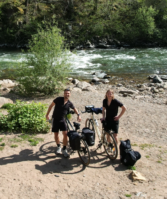 Deux voyageurs à vélo en tenue de sport noire posant avec leurs vélos chargés de sacs près d'une rivière entourée de verdure dans le Pays Basque.
Séjours à vélo clé en main de The Family Bike.