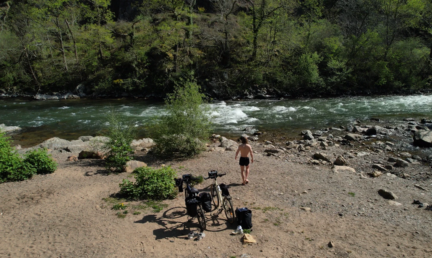 Un voyageur à vélo marche pieds nus vers une rivière au bord d'un chemin de sable avec deux vélos stationnés à proximité.
Séjours à vélo clé en main de The Family Bike.