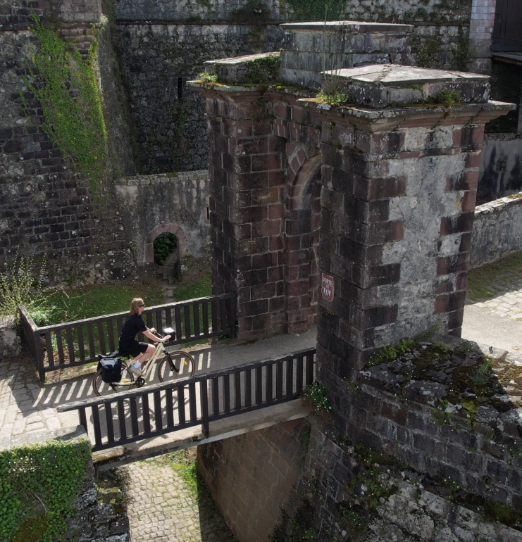 Personne à vélo traversant un petit pont en bois à Saint Jean Pied de Port devant une ancienne fortification en pierre couverte de mousse et de végétation.
Séjours à vélo clé en main de The Family Bike.